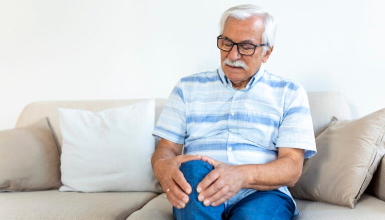Elderly man sitting on a sofa at home and touching his painful knee. people, health care and problem concept - unhappy senior man suffering from knee ache at home