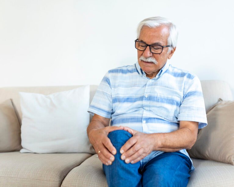 Elderly man sitting on a sofa at home and touching his painful knee. people, health care and problem concept - unhappy senior man suffering from knee ache at home